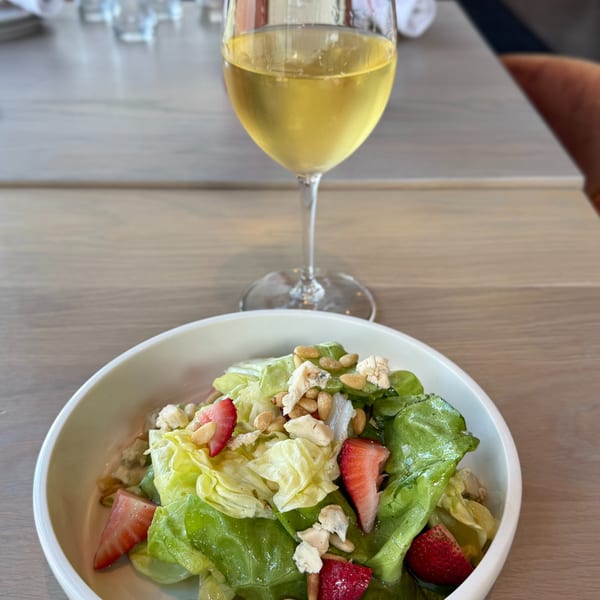 A fresh green salad with strawberries, pine nuts, and cheese, next to a glass of white wine.