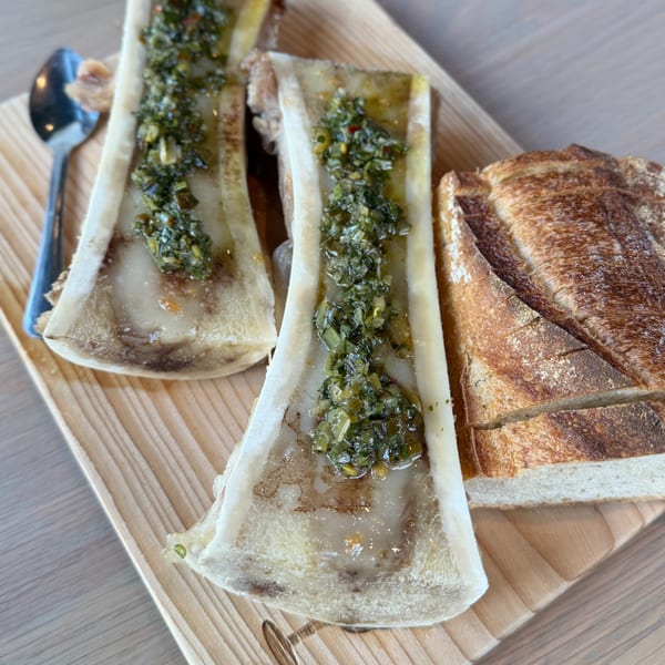 Two bone marrow halves with green herb sauce and sliced bread on a wooden board.