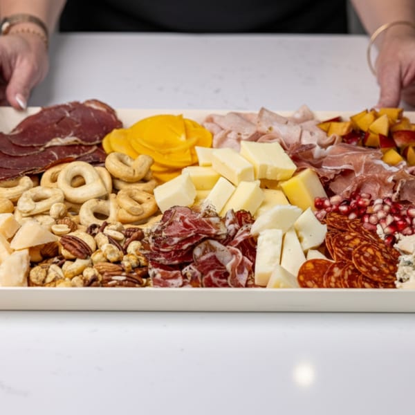 Hands presenting a Crush'd charcuterie board with meats, cheeses, nuts, crackers, and fruit.