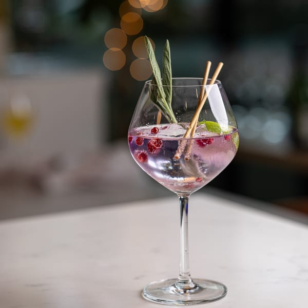 A purple cocktail with cranberries, herbs, and two straws in a large wine glass on a white table.