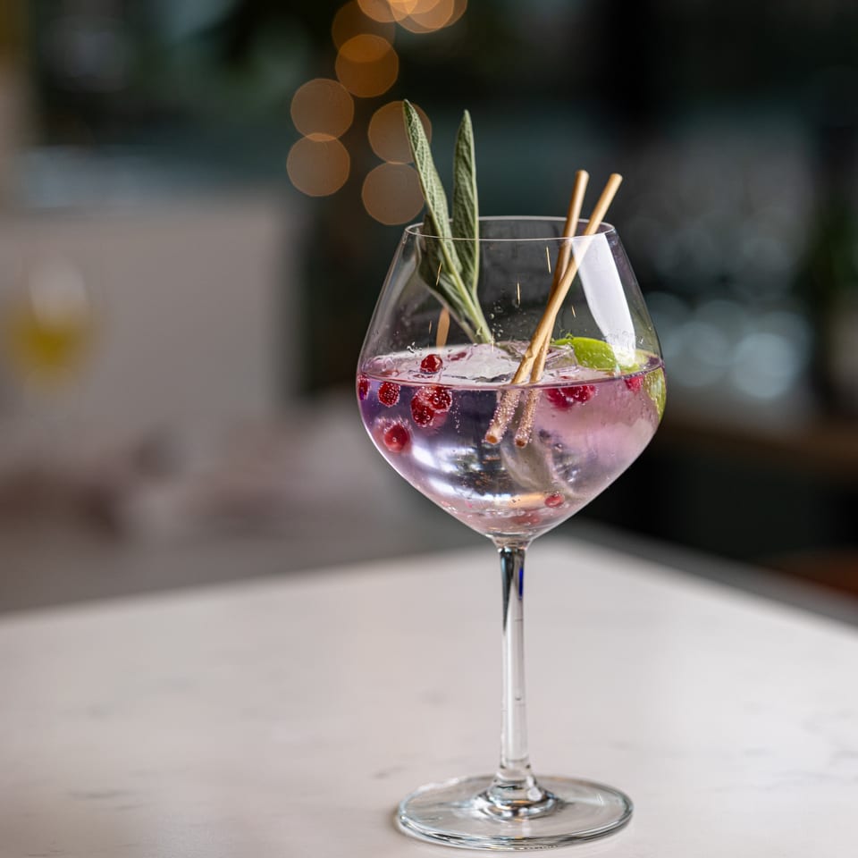 A purple cocktail with cranberries, herbs, and two straws in a large wine glass on a white table.