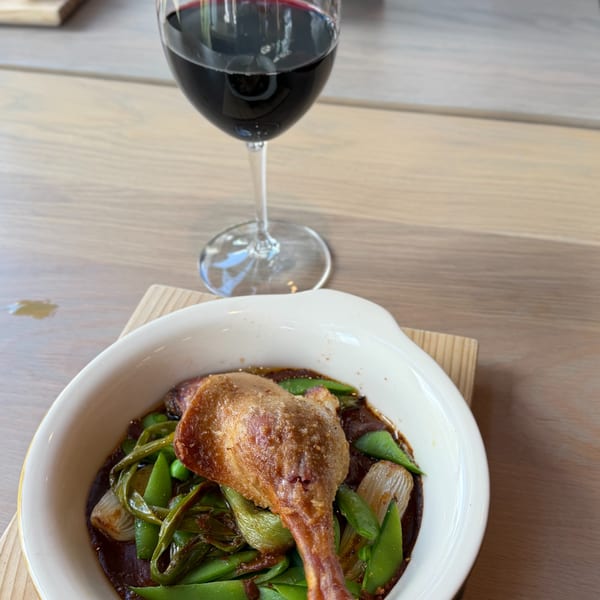A crispy duck leg confit with snap peas and pearl onions in a rich sauce, alongside a glass of red wine.
