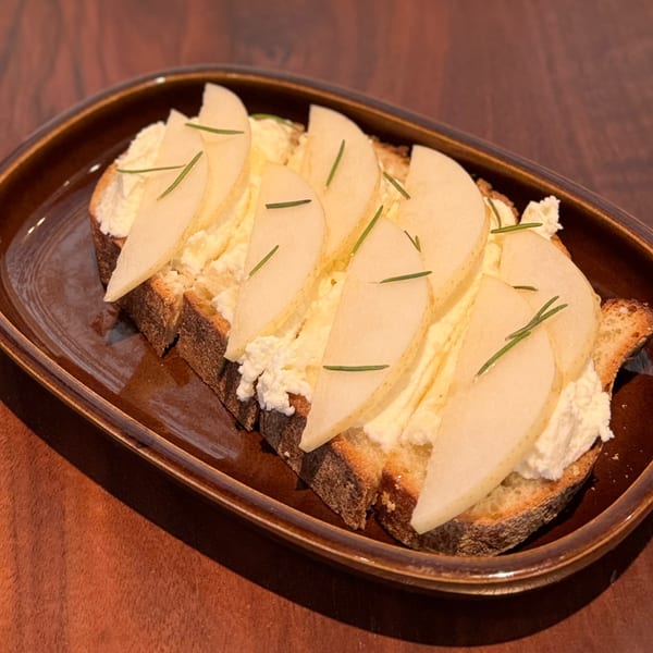 Toasted bread with creamy cheese, thinly sliced pears, and rosemary sprigs on a brown plate.