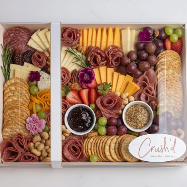 A large charcuterie board in a clear box, filled with meats, cheeses, crackers, fruits, and nuts.
