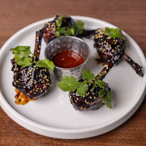 Four glazed chicken lollipops with sesame seeds and cilantro, served with a side of dipping sauce.
