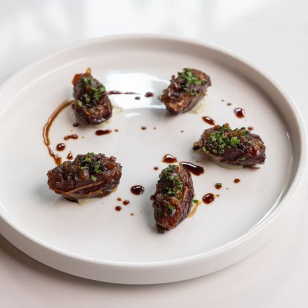 Five dates stuffed with meat, drizzled with sauce and garnished with chives on a white plate.