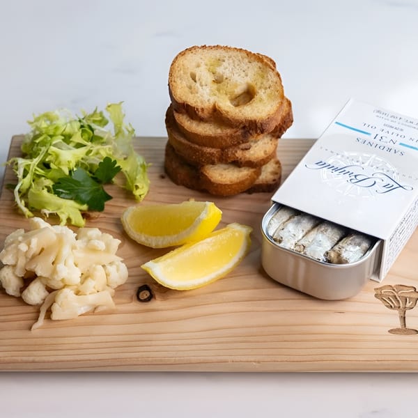 Sardines in a tin with toasted bread, lemon wedges, pickled cauliflower, and lettuce on a wooden board.