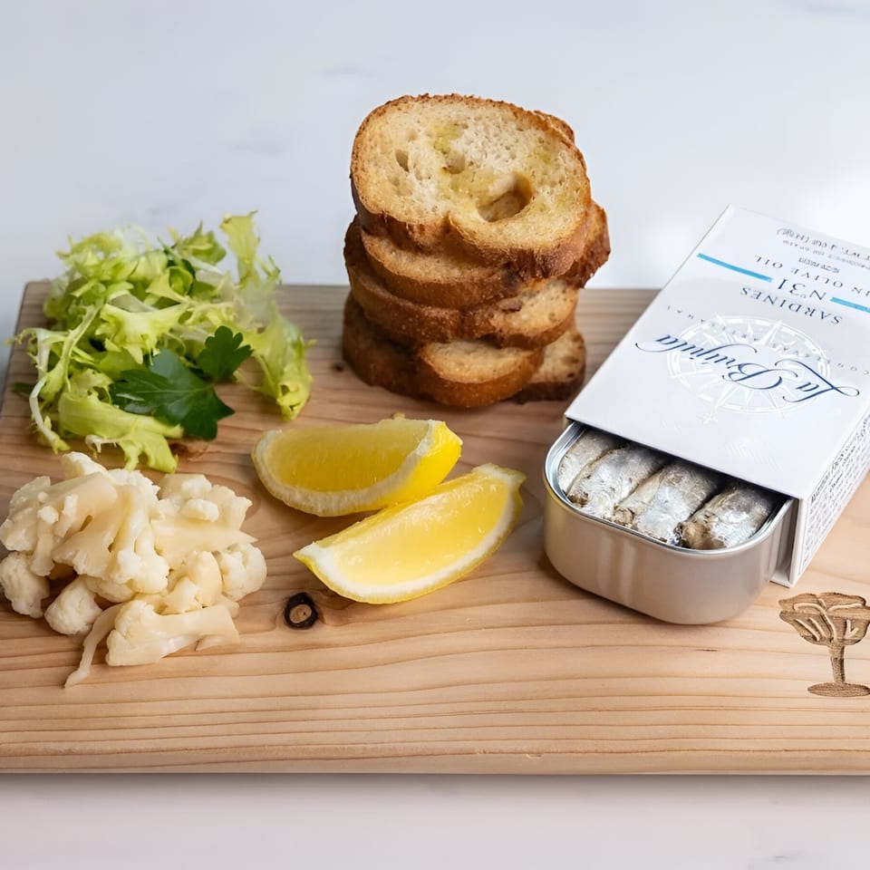 Sardines in a tin with toasted bread, lemon wedges, pickled cauliflower, and lettuce on a wooden board.