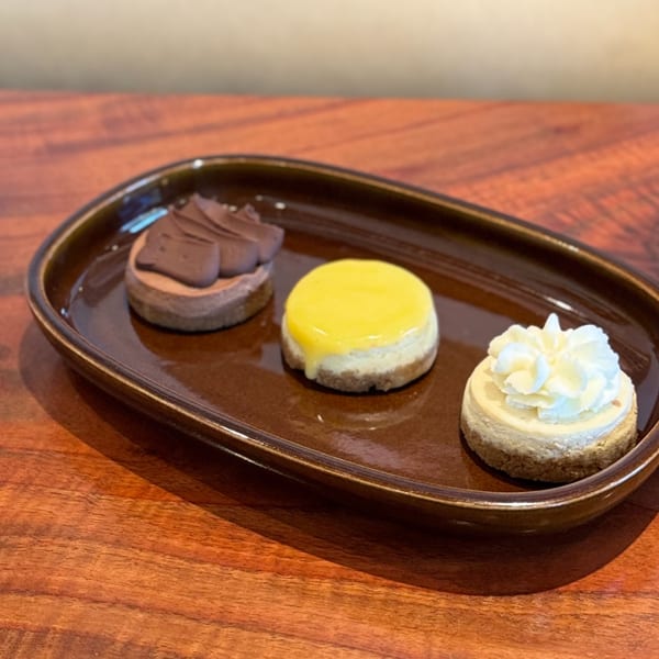 Three mini cheesecakes on a brown plate: chocolate, lemon, and classic with whipped cream.