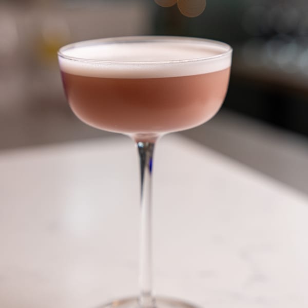 A coupe glass filled with a pinkish-brown cocktail topped with white foam on a white surface.