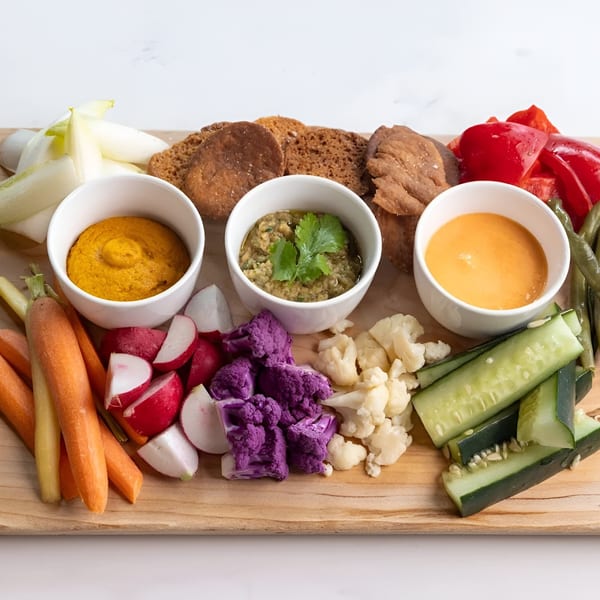 Colorful crudités board with three dips, various vegetables, and toasted bread on a wooden board.