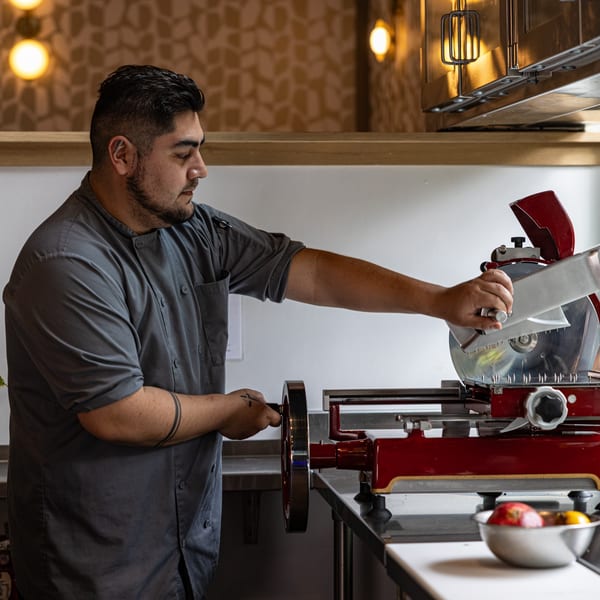 A chef in a gray uniform operates a red meat slicer in a modern kitchen at Crush'd Wine Bar Kitchen.
