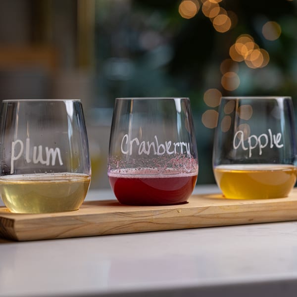 Three wine glasses on a wooden board, labeled Plum, Cranberry, and Apple, filled with different colored drinks.